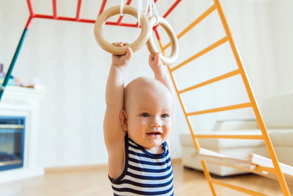 Cute baby performs gymnastic exercises on a wooden home sports complex stairs and rings.